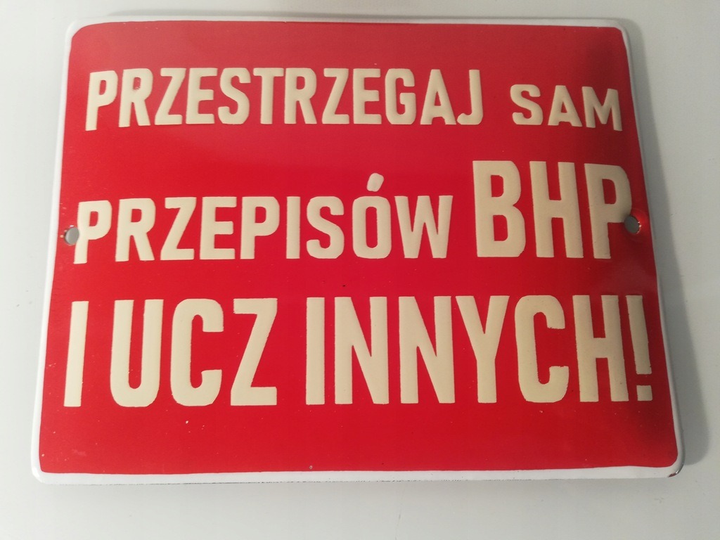 Czerwona tablica informacyjna z napisem 'PRZESTRZEGAJ SAM PRZEPISÓW BHP I UCZ INNYCH!' w kolorze kremowym, przymocowana dwoma nitami.