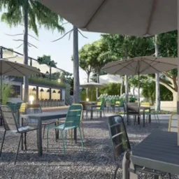 Zewnętrzna przestrzeń restauracji z miejscami siedzącymi pod parasolami, otoczona drzewami i roślinnością, z widocznym budynkiem w tle.