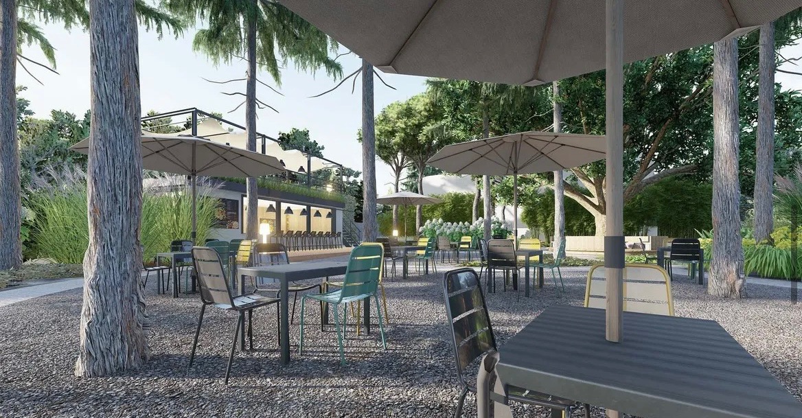 Zewnętrzna przestrzeń restauracji z miejscami siedzącymi pod parasolami, otoczona drzewami i roślinnością, z widocznym budynkiem w tle.