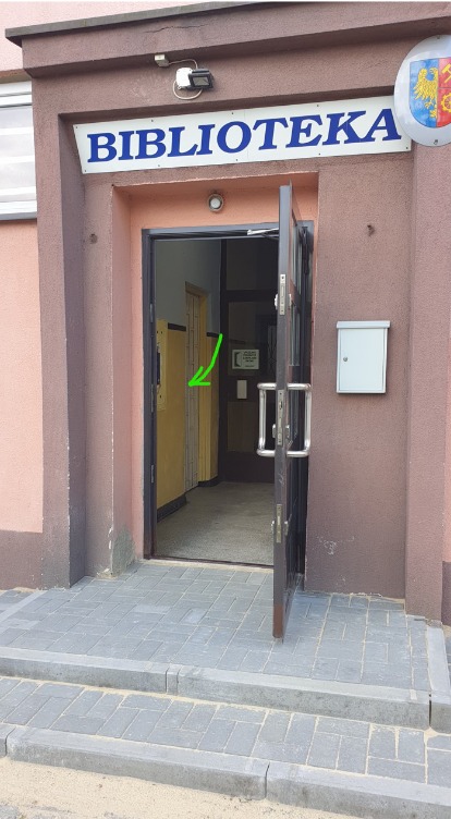 Wejście do biblioteki w Świętochłowicach z otwartymi drzwiami i widoczną kratą zabezpieczającą wewnątrz.