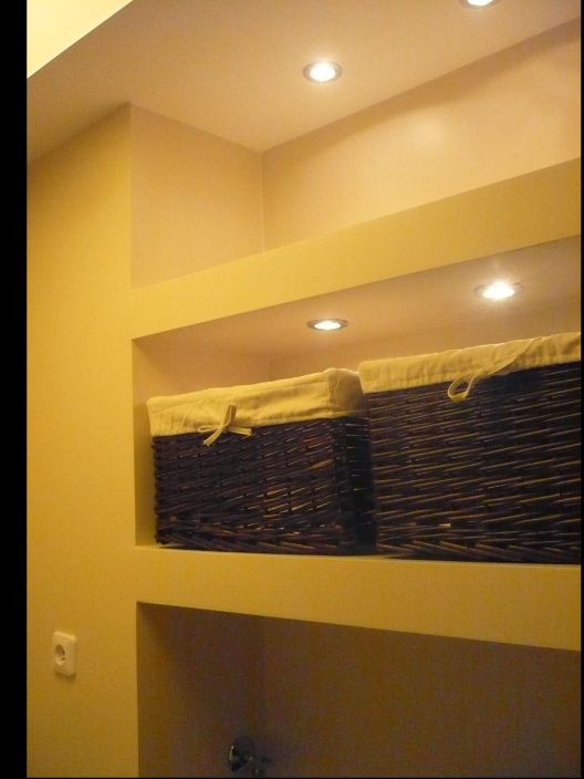 Wnęka w ścianie z dwiema wiklinowymi skrzynkami z materiałowymi wkładami pod sufitem z oświetleniem punktowym, widoczny fragment instalacji elektrycznej.