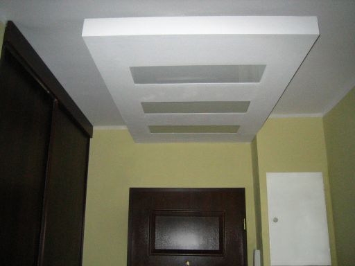 Biały sufit podwieszany z trzema prostokątnymi wnękami w pomieszczeniu o żółtych ścianach, widoczna część ciemnobrązowej szafy przesuwnej i drzwi po lewej stronie.