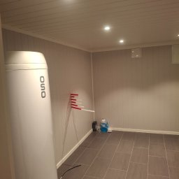 Mikołaj Kulig - Wnętrze pomieszczenia z grzejnikiem elektrycznym OSO na tle ściany z boazerią, szara podłoga z płytek, widoczne akcesoria montażowe i oświetlenie sufitowe