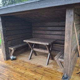 Mikołaj Kulig - Zadaszona konstrukcja z ciemnego drewna z ławkami i stołem na jasnym drewnianym tarasie, widoczne dekoracyjne lampiony i świece w szklanych pojemnikach.