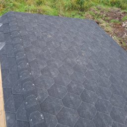 Mikołaj Kulig - Fragment dachu pokryty ciemną, heksagonalną papą bitumiczną, widoczny z góry, z fragmentem drewnianego podestu obok i ziemią z roślinnością w tle.