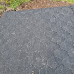 Mikołaj Kulig - Fragment dachu pokryty szarym gontem bitumicznym o heksagonalnym kształcie, mocowanym na gwoździe, z widocznym fragmentem gruntu i roślinności w tle.