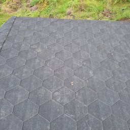 Mikołaj Kulig - Widok z góry na fragment dachu pokrytego czarnym gontem bitumicznym o heksagonalnym kształcie, z widocznymi łączeniami i fragmentem zieleni w tle.