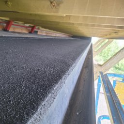 Mikołaj Kulig - Widok z dołu na fragment dachu pokrytego czarną papą, z widocznymi elementami konstrukcji drewnianej i fragmentem podnośnika koszowego z boku.