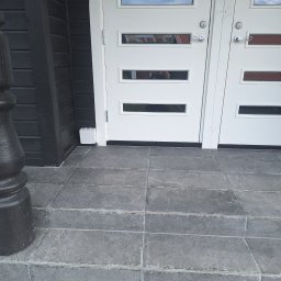 Mikołaj Kulig - Szare płytki schodowe prowadzące do białych drzwi wejściowych z poziomymi przeszkleniami, obok czarna drewniana elewacja budynku z dekoracyjnym filarem.