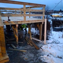 Mikołaj Kulig - Budowa drewnianego tarasu w zimowej scenerii: widoczne elementy konstrukcyjne, narzędzia i pracownik w odblaskowej kamizelce przy sprzątaniu placu budowy.