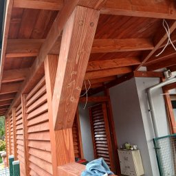 Mikołaj Kulig - Zadaszenie tarasu z drewna o ciepłym odcieniu, z widoczną konstrukcją wspornikową i lamelową ścianą. Na pierwszym planie fragment balustrady i niebieska ściereczka.