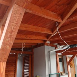 Mikołaj Kulig - Drewniana konstrukcja z belek i desek w ciepłym odcieniu brązu, widok z dołu, ukazujący detale połączeń i fakturę drewna, z fragmentem budynku w tle.