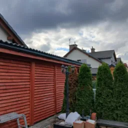 Czerwona, drewniana zabudowa z numerem domu 208, widoczna drabina, lampa i tuje. W tle domy pod pochmurnym niebem.