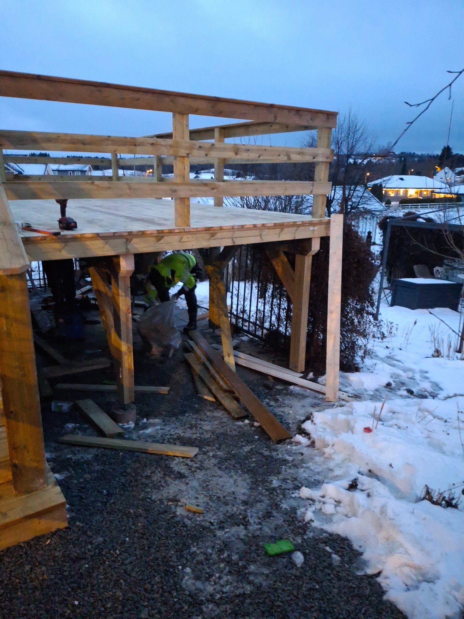 Budowa drewnianego tarasu w zimowej scenerii: widoczne elementy konstrukcyjne, narzędzia i pracownik w odblaskowej kamizelce przy sprzątaniu placu budowy.