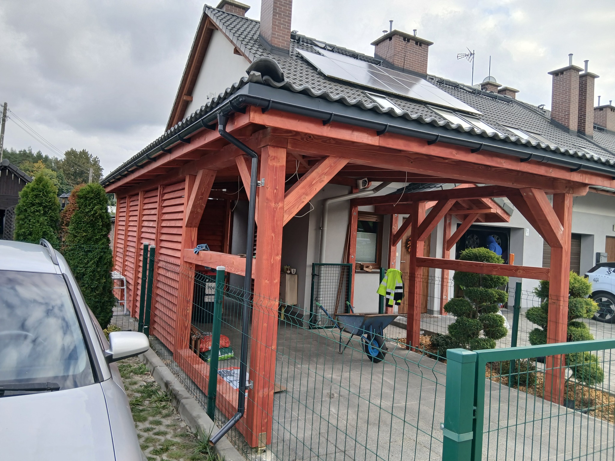 Konstrukcja drewniana przy domu z panelami słonecznymi na dachu, widoczny wózek i ogrodzenie. Częściowo widoczny samochód.