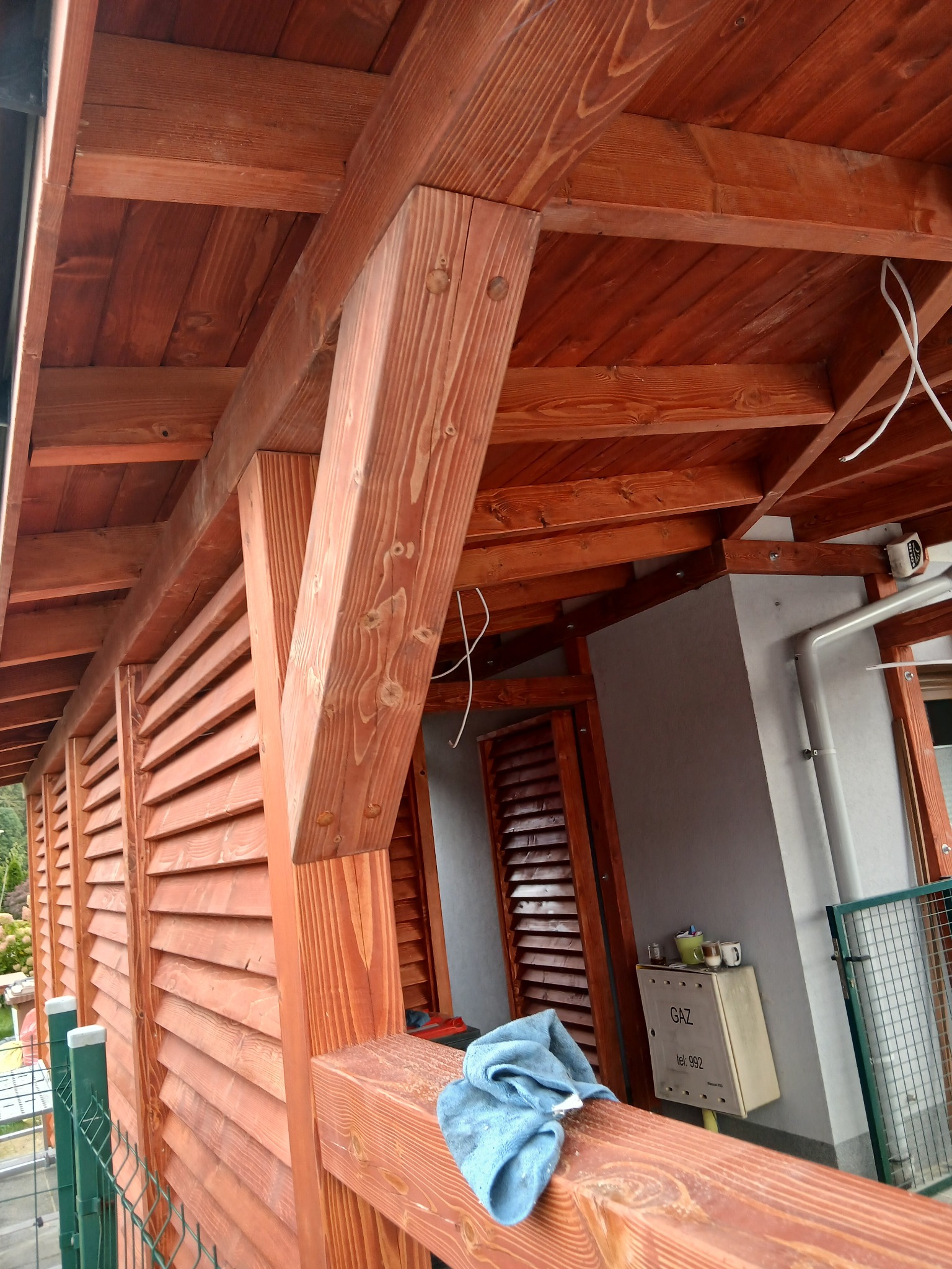 Zadaszenie tarasu z drewna o ciepłym odcieniu, z widoczną konstrukcją wspornikową i lamelową ścianą. Na pierwszym planie fragment balustrady i niebieska ściereczka.