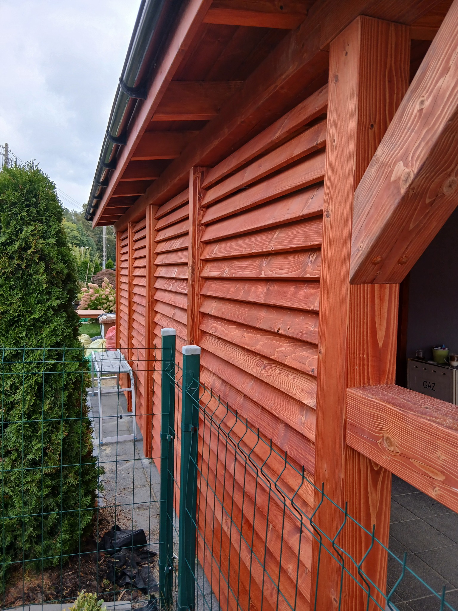 Drewniana konstrukcja z lamelami w kolorze pomarańczowym, widoczna z zewnątrz, z fragmentem zielonego ogrodzenia i roślinności w tle. Detal wykończenia i montażu.