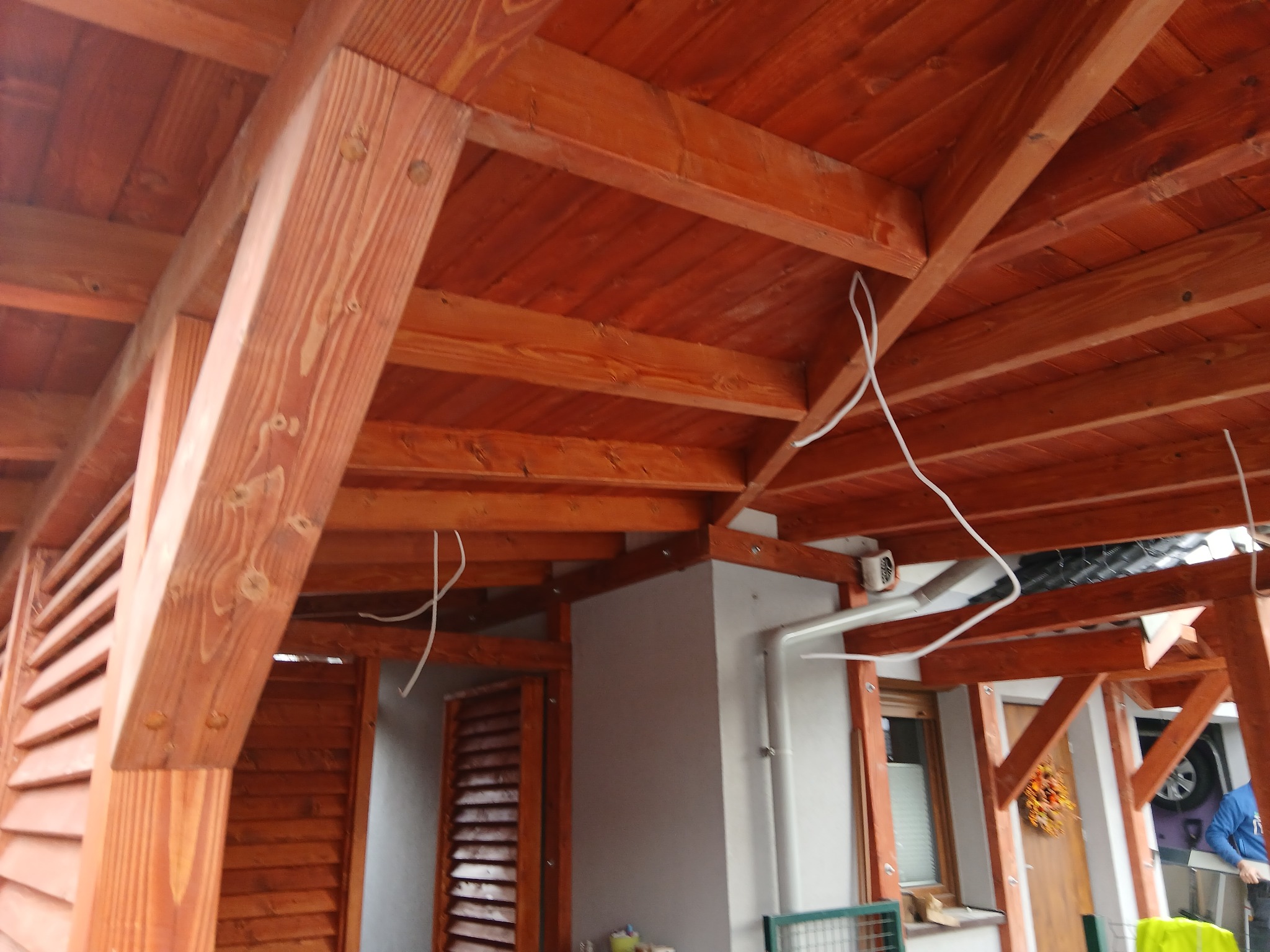 Drewniana konstrukcja z belek i desek w ciepłym odcieniu brązu, widok z dołu, ukazujący detale połączeń i fakturę drewna, z fragmentem budynku w tle.