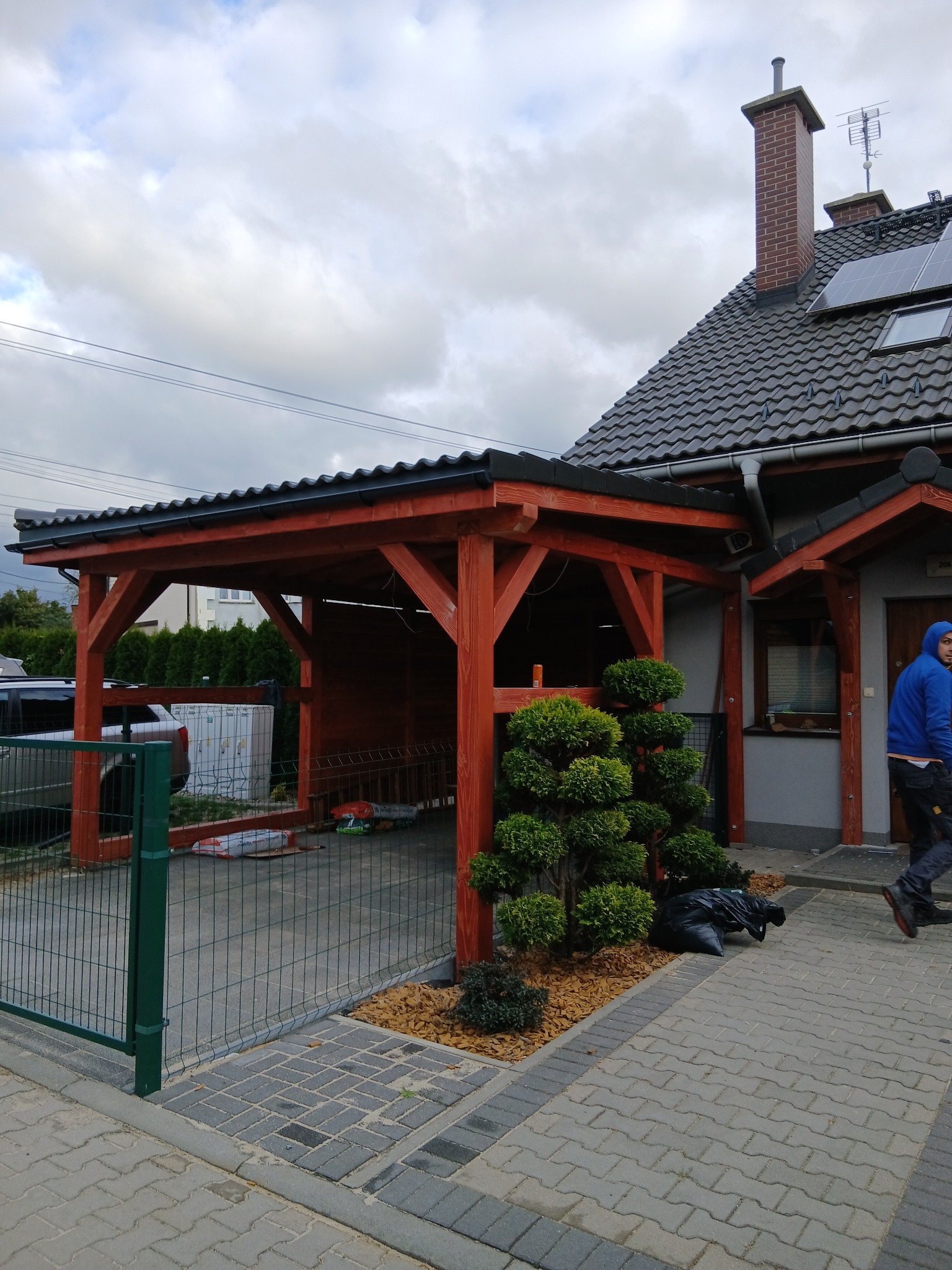 Drewniana wiata samochodowa z czarnym dachem, przylegająca do domu z szarą elewacją i brązowymi elementami. Na pierwszym planie zielona brama i ozdobne krzewy.