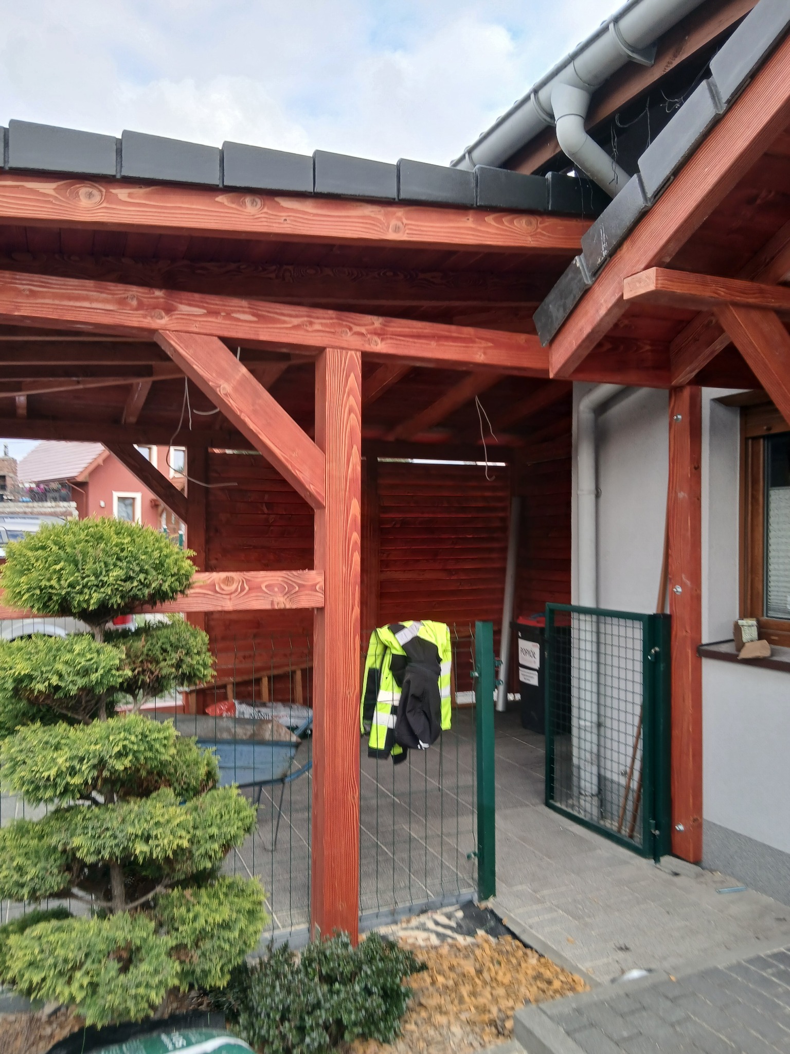 Częściowy widok drewnianej konstrukcji z czarnym dachem i rynną, zielona brama i drzewko bonsai na pierwszym planie, w tle widoczny dom.
