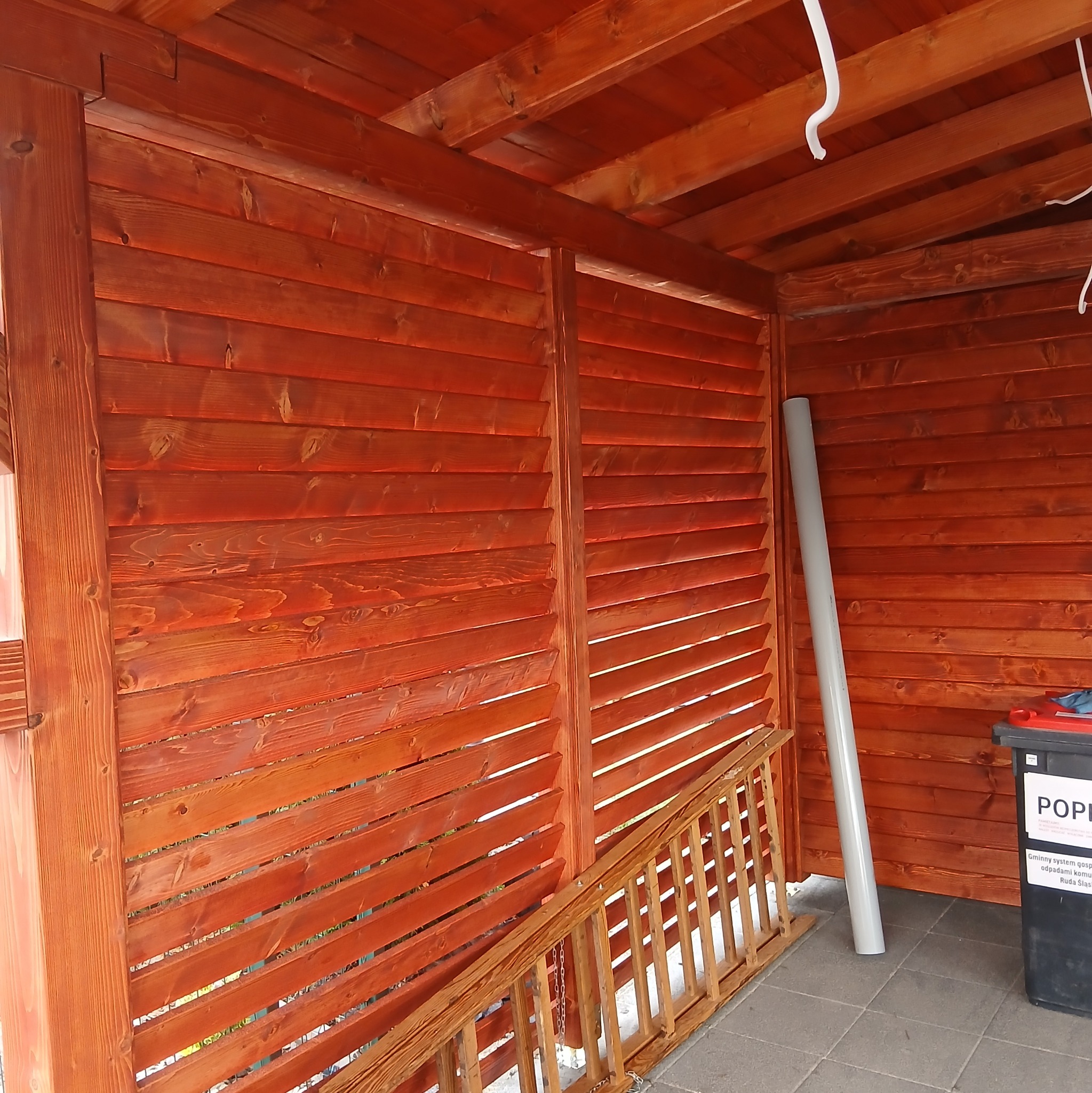 Drewniana konstrukcja z ażurową ścianą, pomalowana na czerwono-brązowy kolor. Widoczne elementy konstrukcyjne dachu i fragment podłogi z kostki brukowej.
