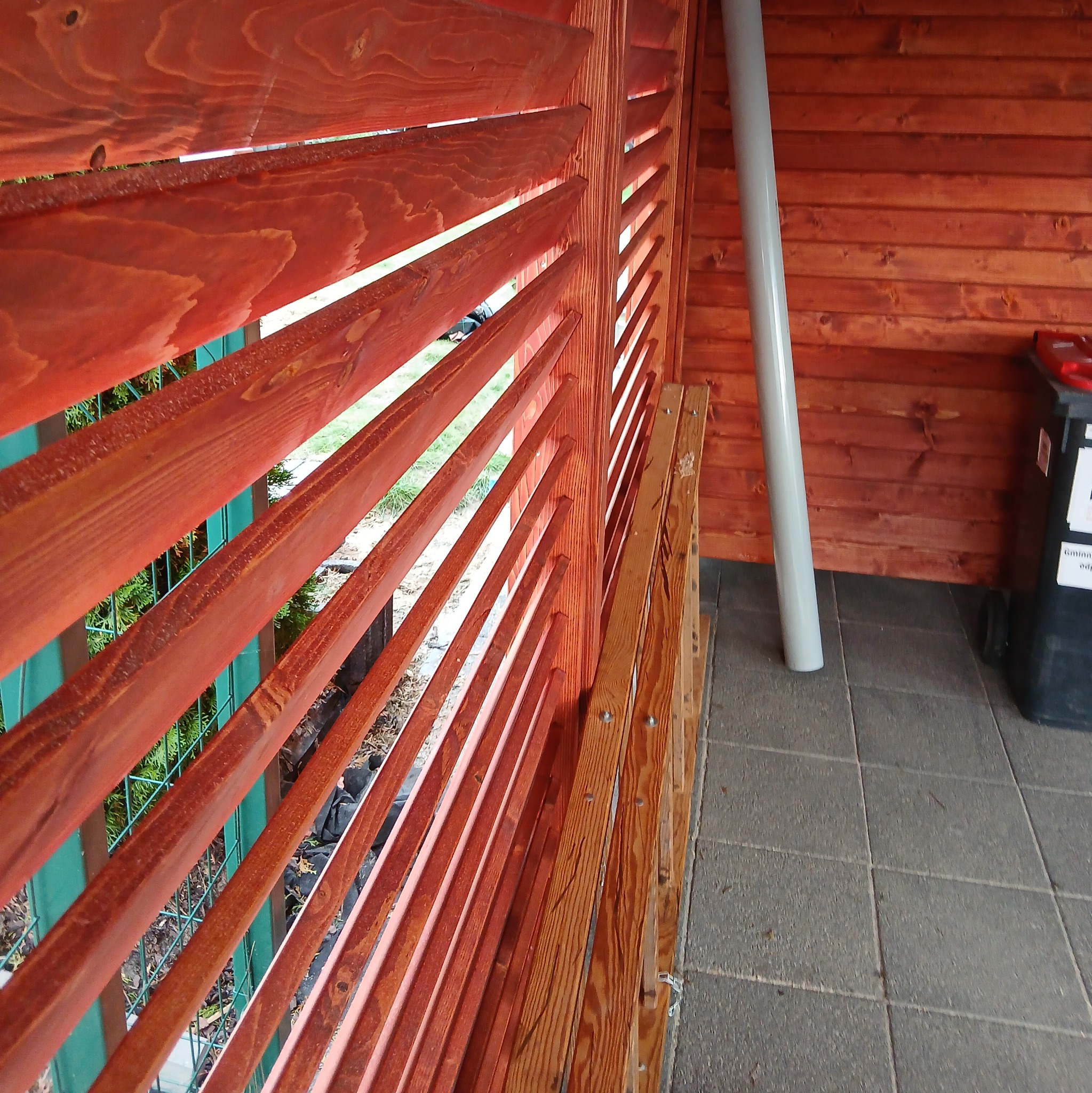 Drewniana konstrukcja z listew w ciepłym odcieniu brązu, tworząca przesłonę z widokiem na zieleń. Widoczne detale montażowe i fragment otoczenia z szarymi płytkami.