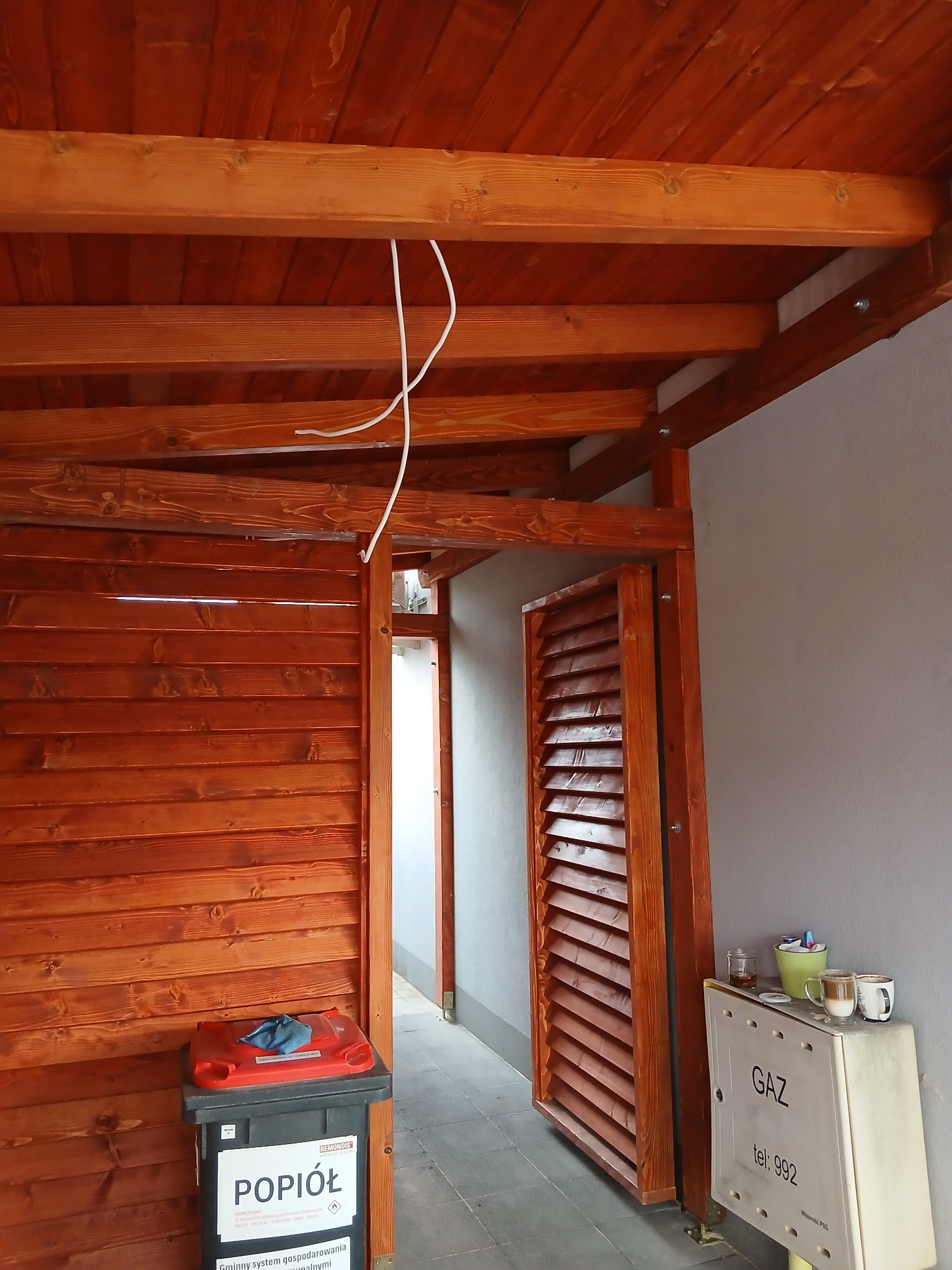 Drewniana konstrukcja z lamelami i podwieszanym sufitem z desek. Widoczne przewody elektryczne, śmietnik i szafka gazowa. Ciepłe oświetlenie podkreśla strukturę drewna.