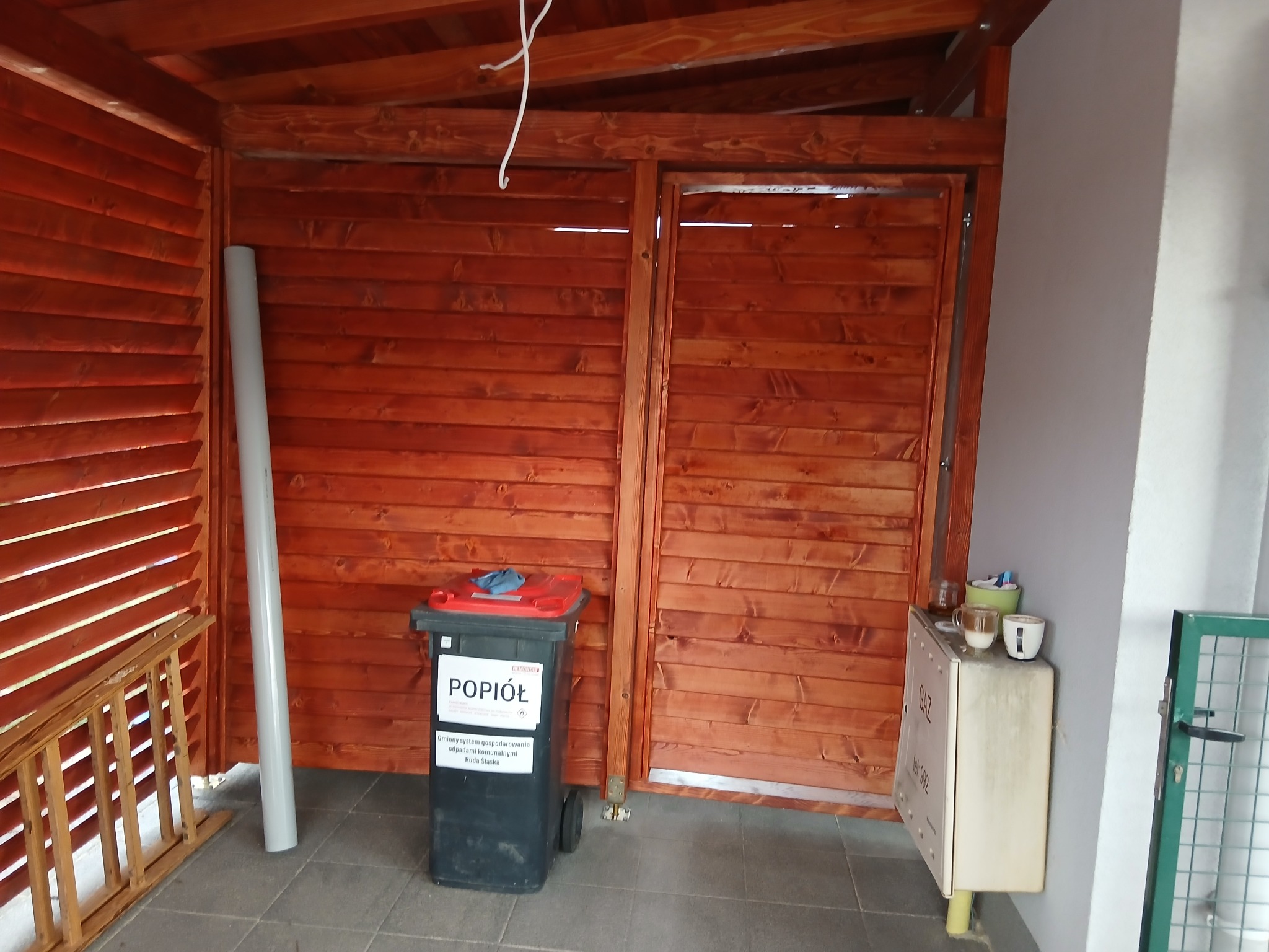 Drewniana konstrukcja z lamelami, prawdopodobnie altana lub zadaszenie, z koszem na popiół i rurą PCV w rogu, ukazana w dziennym świetle.