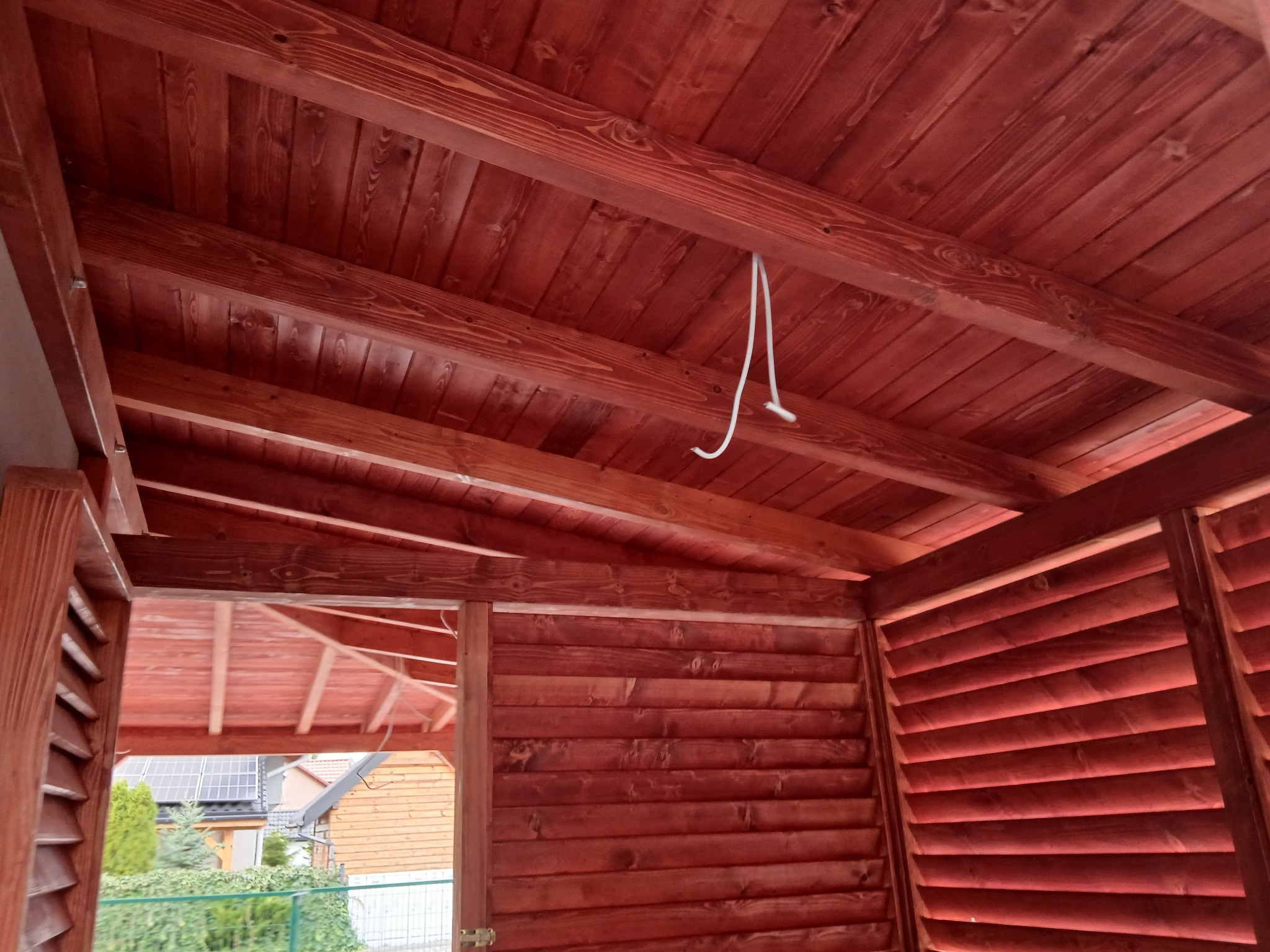 Czerwona drewniana konstrukcja z widocznym dachem z desek i fragmentem ściany z żaluzjami. Widoczny kabel zwisający z dachu. Na zewnątrz zieleń i budynki.
