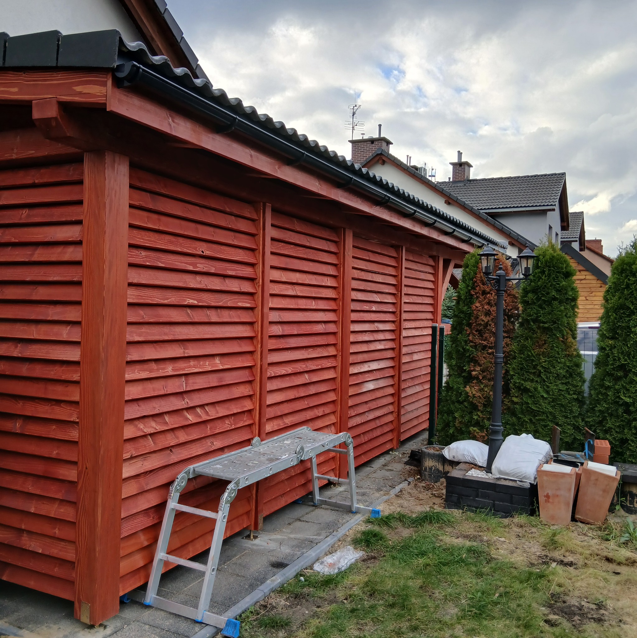 Czerwona, drewniana konstrukcja z lamelami, widoczna z boku, z drabinką i materiałami budowlanymi obok. Tło stanowią domy i pochmurne niebo.