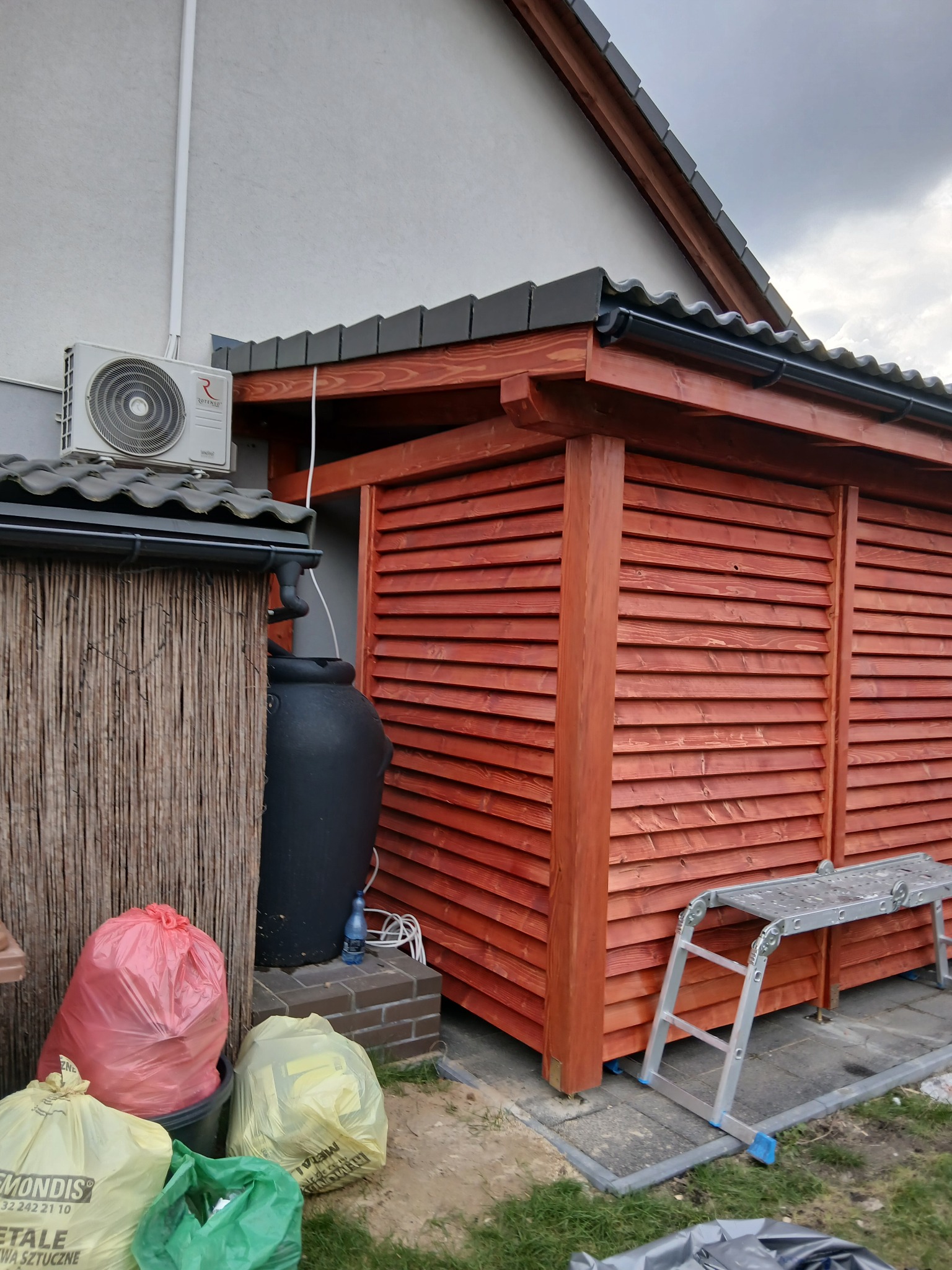 Czerwona, drewniana konstrukcja z lamelami przy ścianie budynku, obok zbiornik na deszczówkę i śmieci. Widoczna drabina i klimatyzator na elewacji.