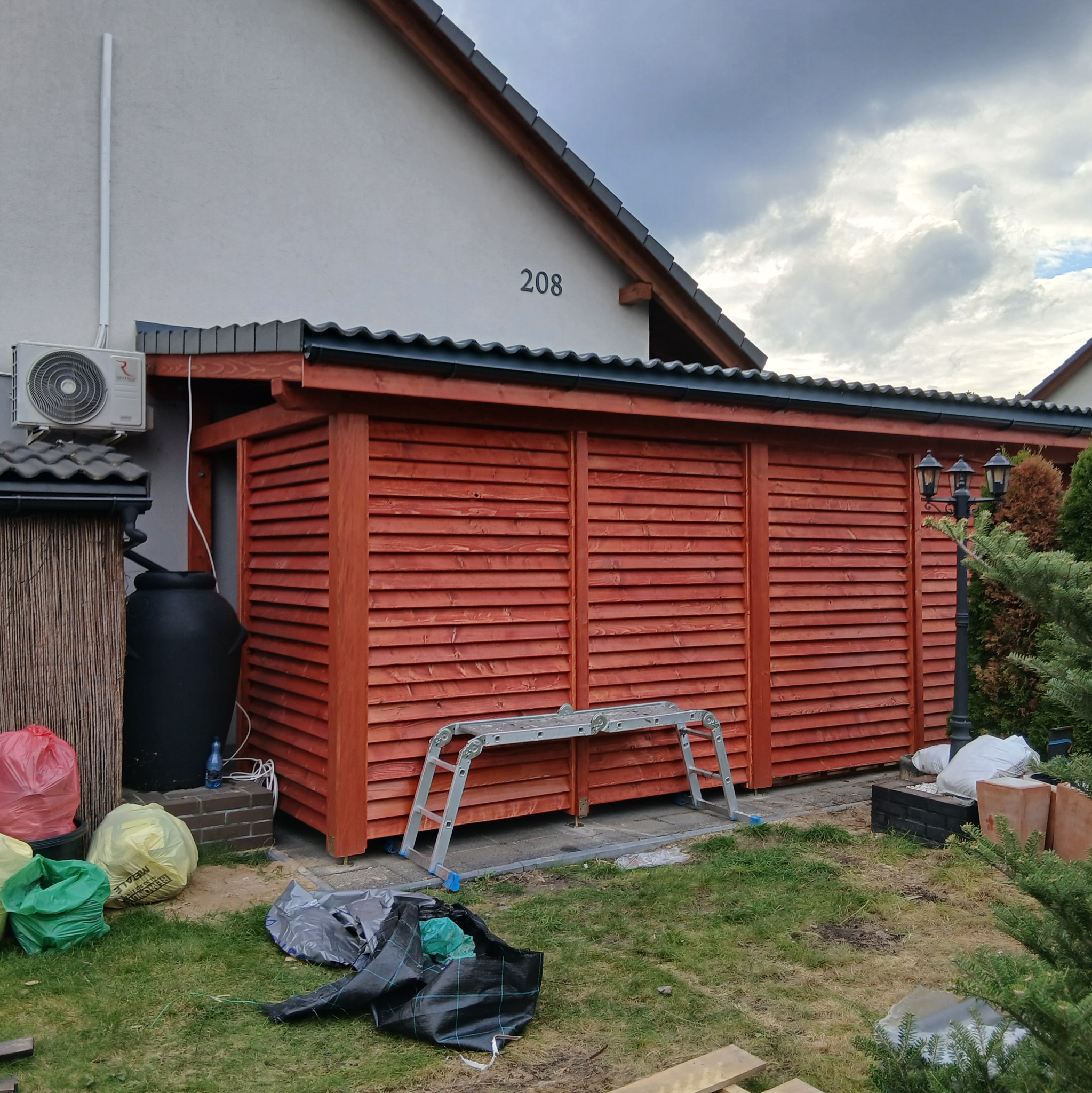 Drewniana, ażurowa zabudowa na posesji z numerem 208, z rozstawioną drabiną i workami w tle. Widoczny fragment domu, lampa ogrodowa i zieleń.