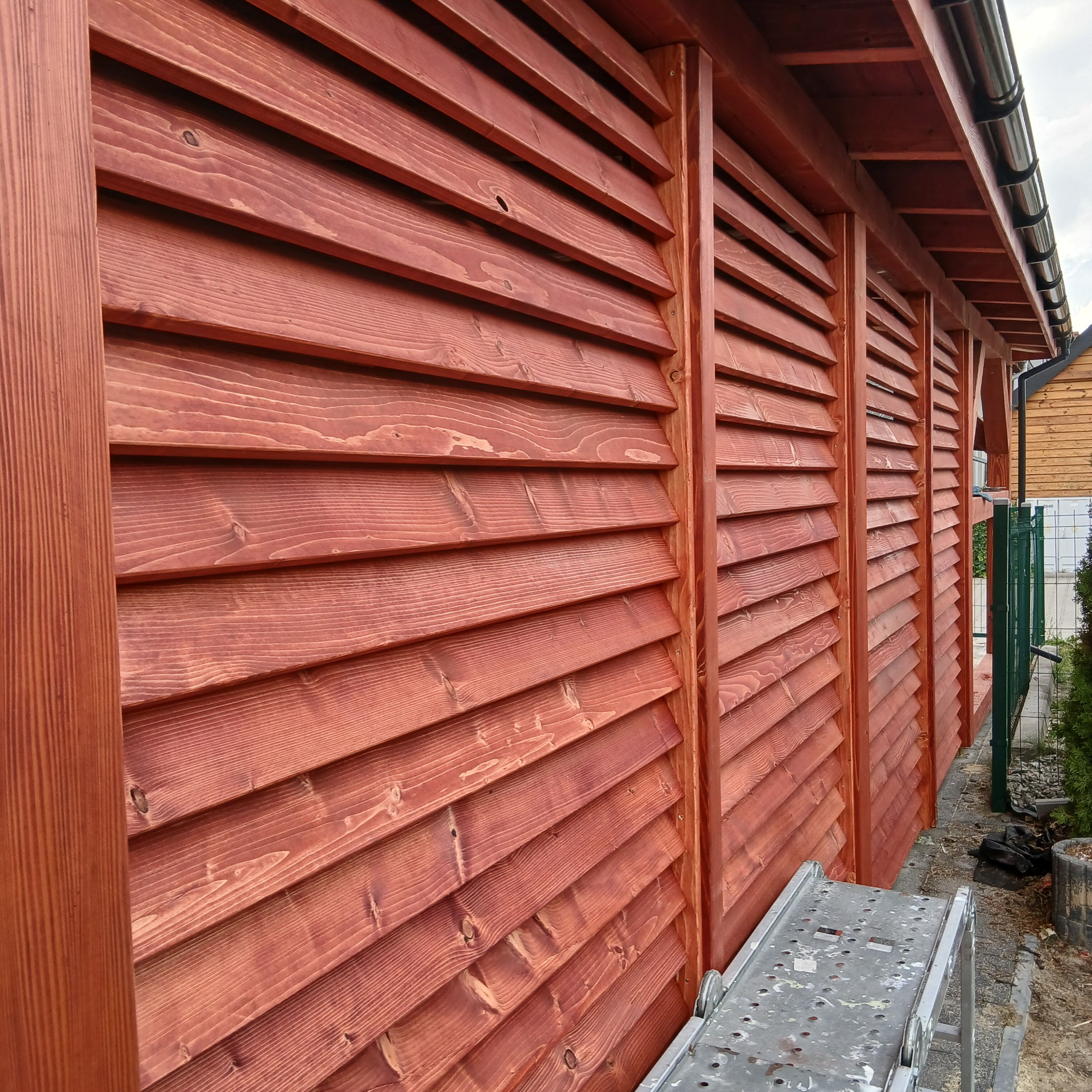 Detale drewnianej konstrukcji z lamelami w odcieniach czerwieni, widoczne z boku, z fragmentem drabiny malarskiej w dolnej części kadru.