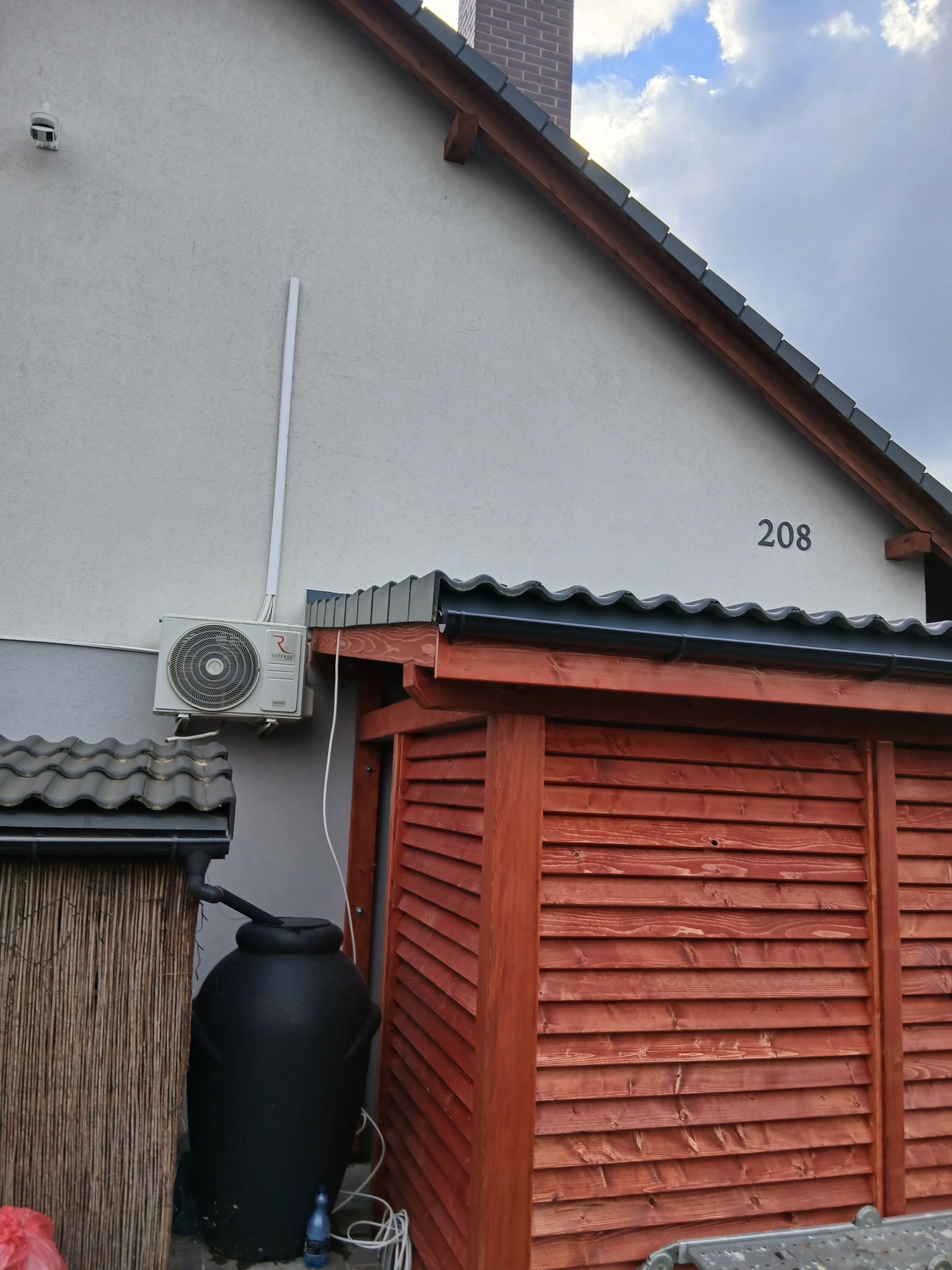 Fragment budynku z numerem 208, drewnianą zabudową w kolorze czerwonym i klimatyzatorem na ścianie. Widoczny również zbiornik na wodę i rynna.