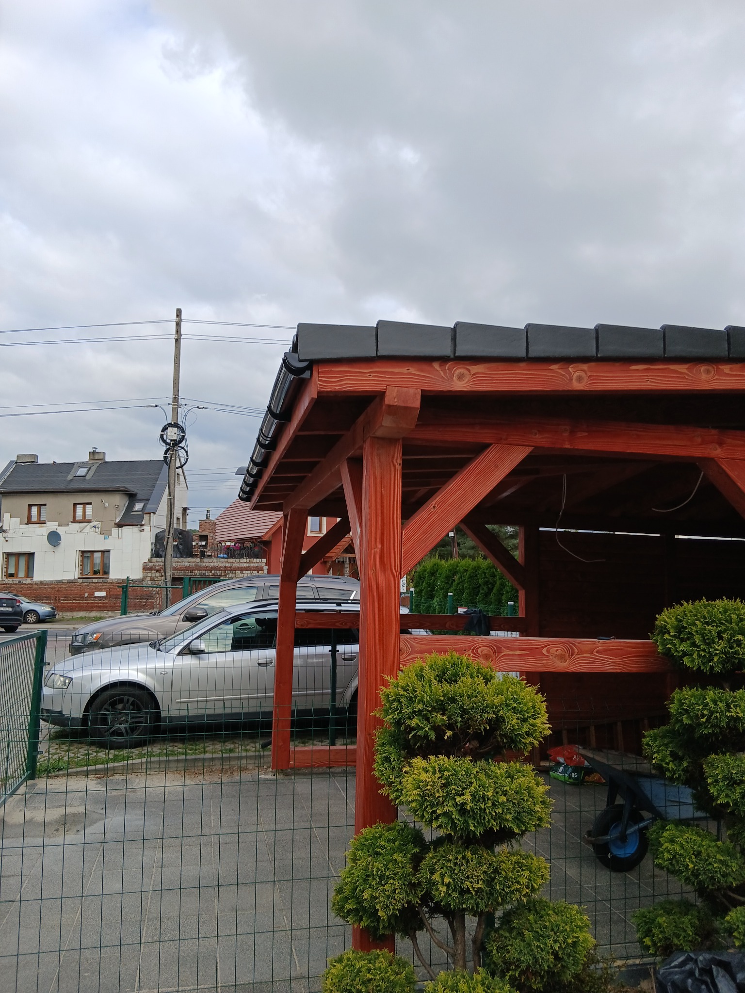 Czerwona, drewniana pergola z czarnym dachem na tle pochmurnego nieba. Obok zaparkowane auta i ozdobne krzewy. Widoczny fragment posesji.