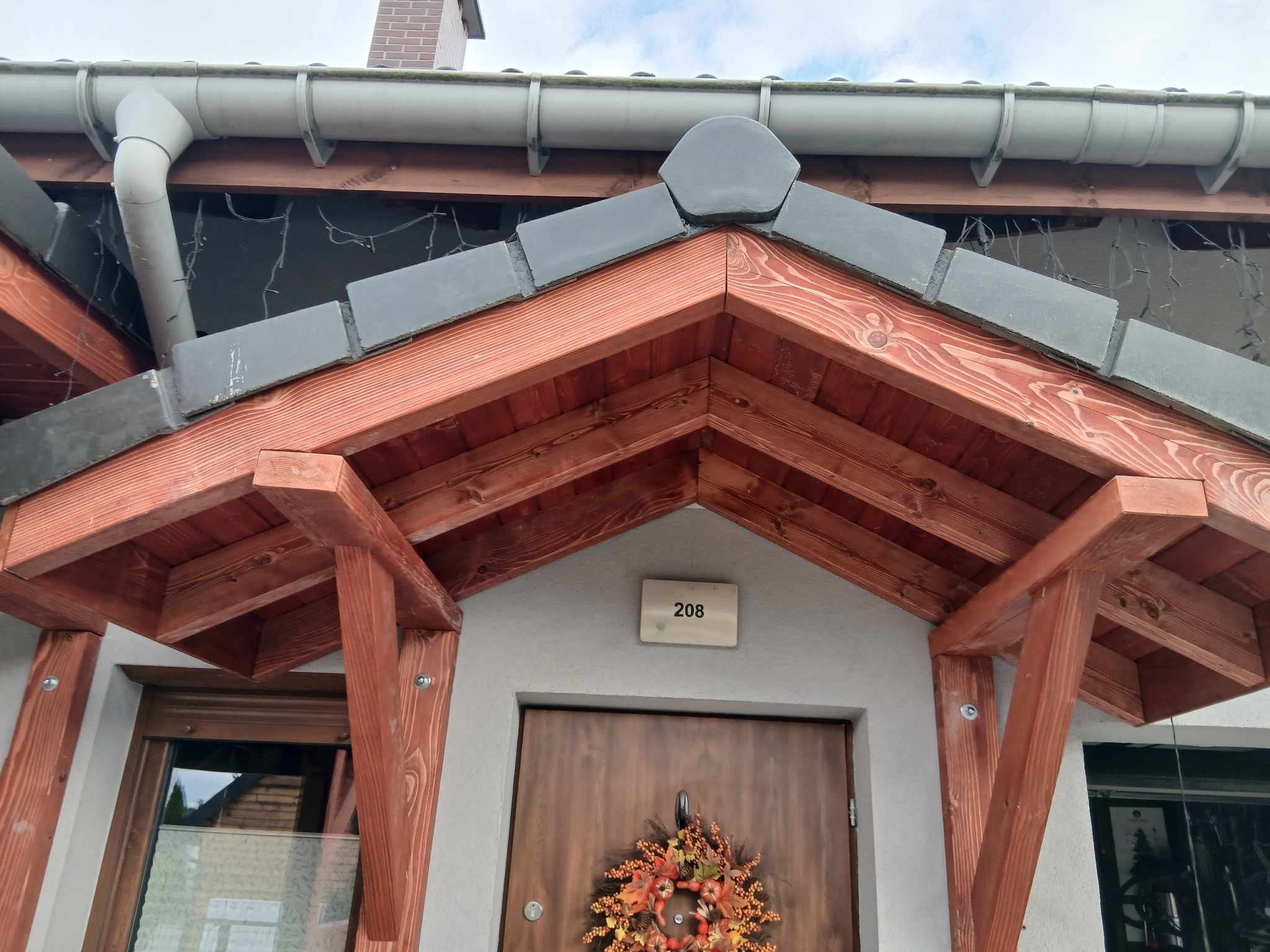 Wejście do domu z drewnianą konstrukcją zadaszenia, szarym dachem i jesiennym wieńcem na brązowych drzwiach. Widoczny numer domu.