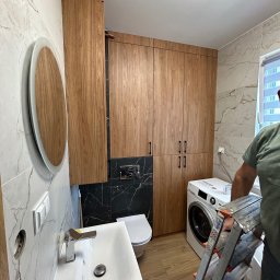 43Remont - Nowoczesna łazienka z drewnianymi szafkami, umywalką nablatową, okrągłym lustrem z podświetleniem LED i pralką, widoczna drabina Krause sugerująca trwające prace wykończeniowe.