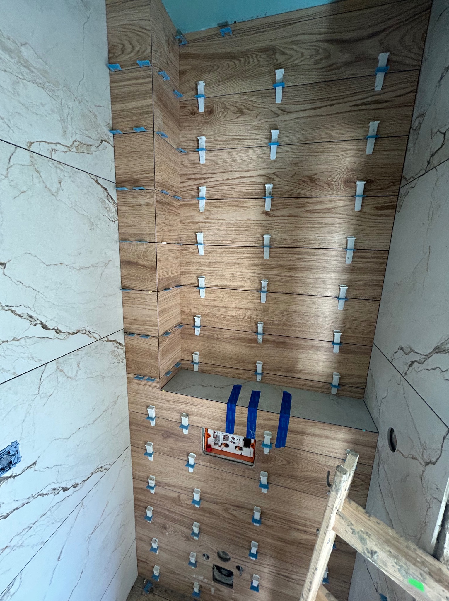 Montaż płytek imitujących drewno w narożniku łazienki, użycie plastikowych krzyżyków dystansowych, widoczna drabina malarska.