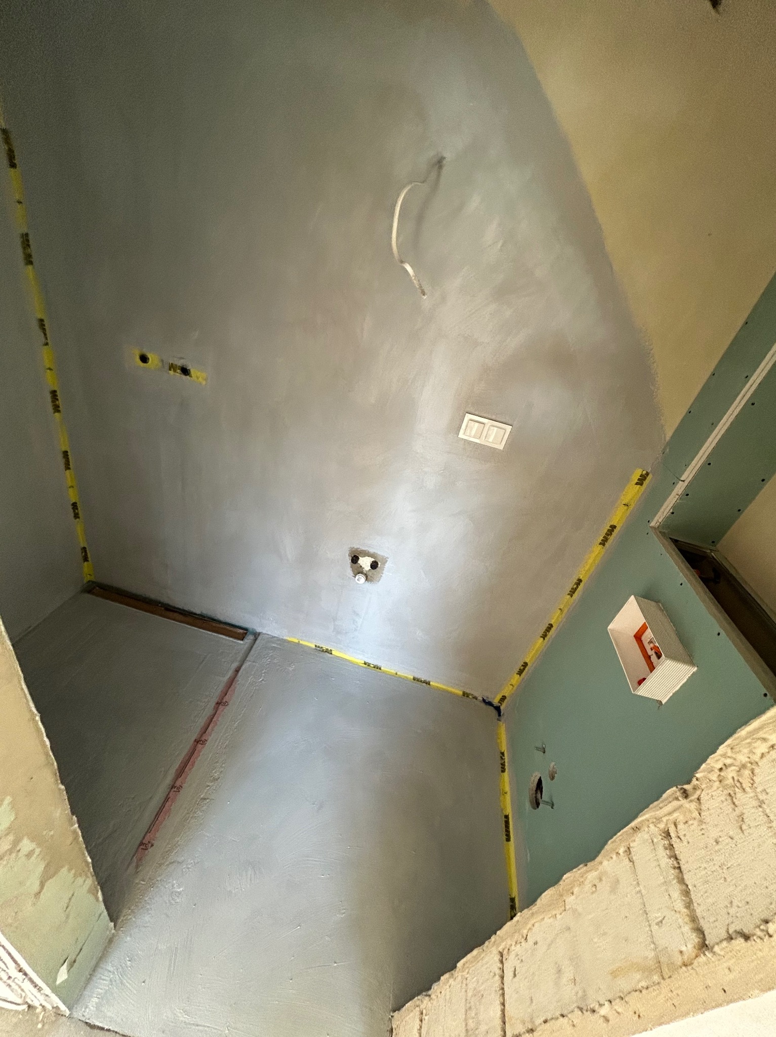 Wnętrze łazienki w trakcie remontu: ściany i sufit pokryte świeżo nałożoną gładzią, zabezpieczone żółtą taśmą malarską wzdłuż krawędzi, widoczne puszki elektryczne i fragment instalacji, płyty...