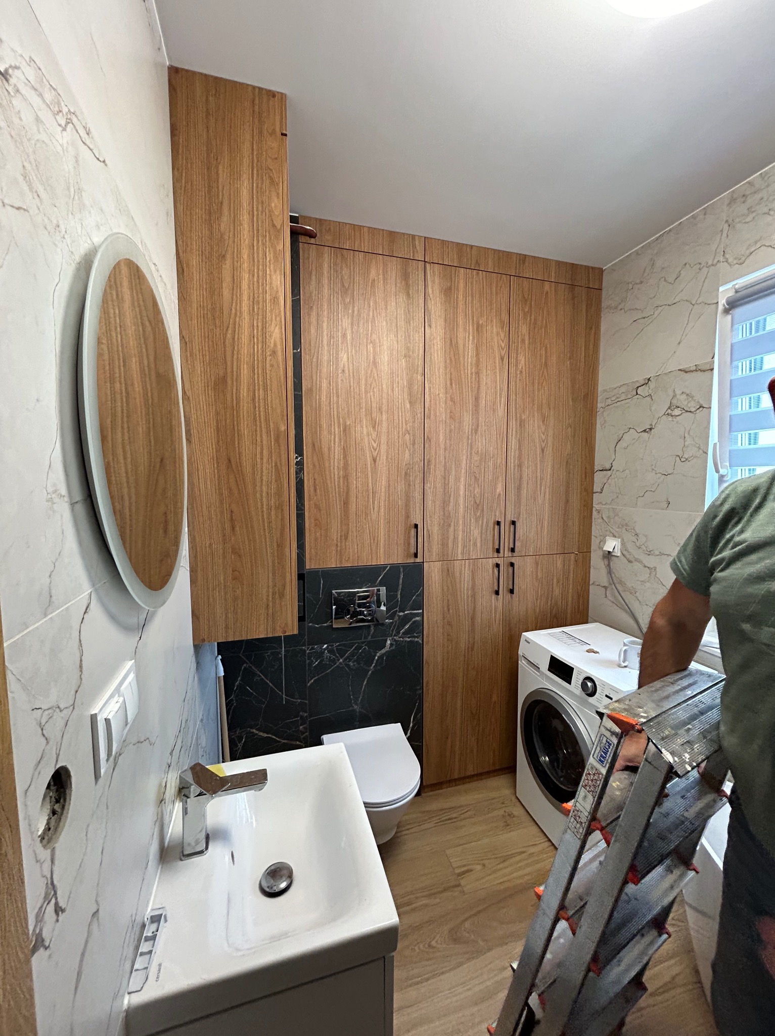 Nowoczesna łazienka z drewnianymi szafkami, umywalką nablatową, okrągłym lustrem z podświetleniem LED i pralką, widoczna drabina Krause sugerująca trwające prace wykończeniowe.