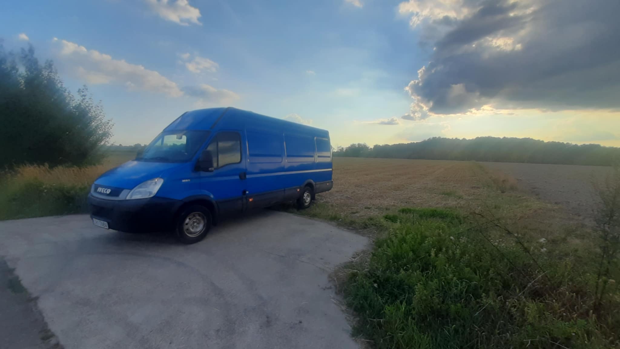 Niebieski bus marki Iveco zaparkowany na betonowej nawierzchni z polami i drzewami w tle, sfotografowany w słoneczny dzień z widocznymi chmurami.