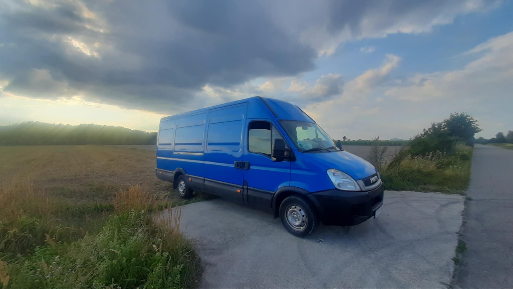 Niebieski bus dostawczy Iveco na betonowej nawierzchni, z polem i zachmurzonym niebem w tle.