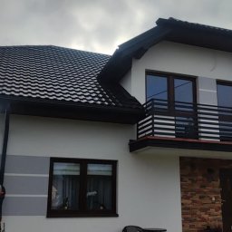 Usługi dekarskie - Pracownik na rusztowaniu montuje rynnę na tle domu z ciemną dachówką i balkonem.