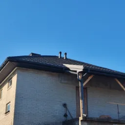 Dom z szarej cegły z nowym, ciemnym dachem z rynnami, widoczne kominy i fragment rusztowania.