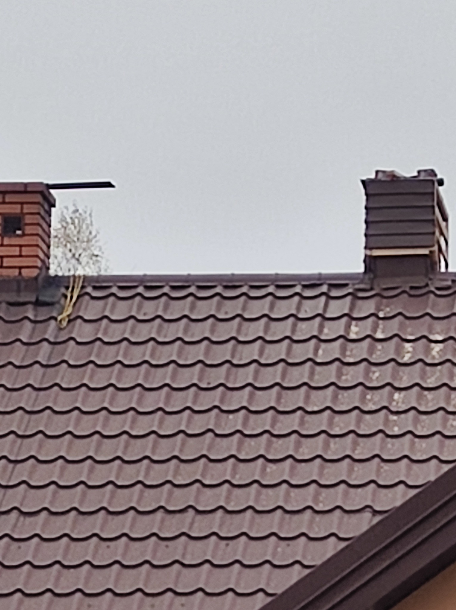 Brązowy dach z metalowej dachówki z dwoma kominami, jednym z cegły i drugim z brązowych paneli, na tle szarego nieba.