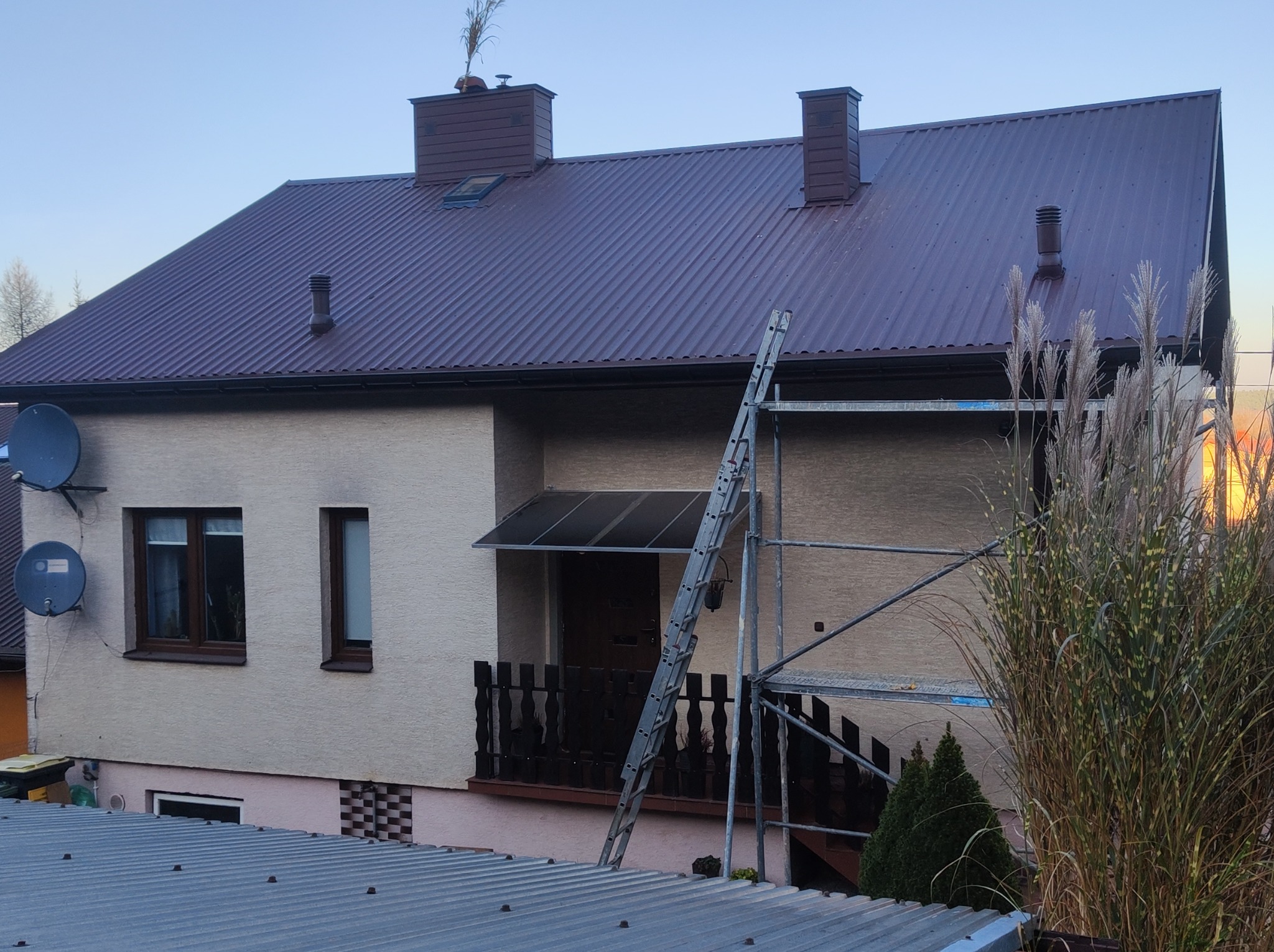 Dom jednorodzinny z ciemnobrązowym dachem z blachy trapezowej, drabina oparta o ścianę, widoczne rusztowanie i ozdobne trawy przed budynkiem.