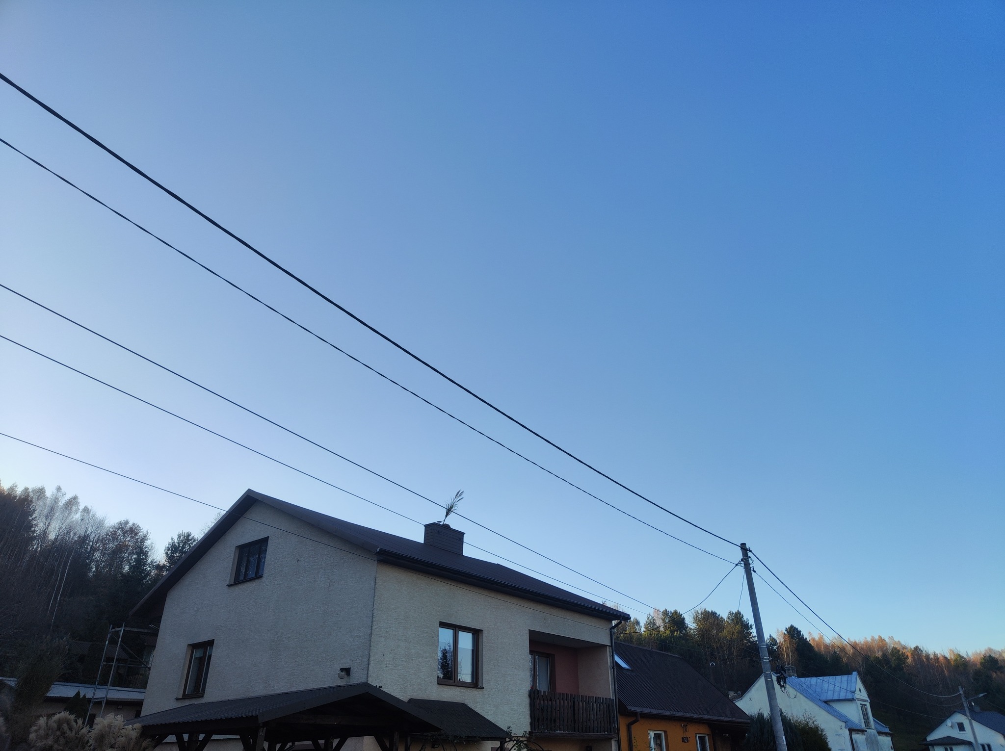 Dwupiętrowy dom z ciemnym dachem i jasną elewacją, widoczny z dołu pod kątem, z drzewami w tle i liniami energetycznymi przecinającymi niebo.