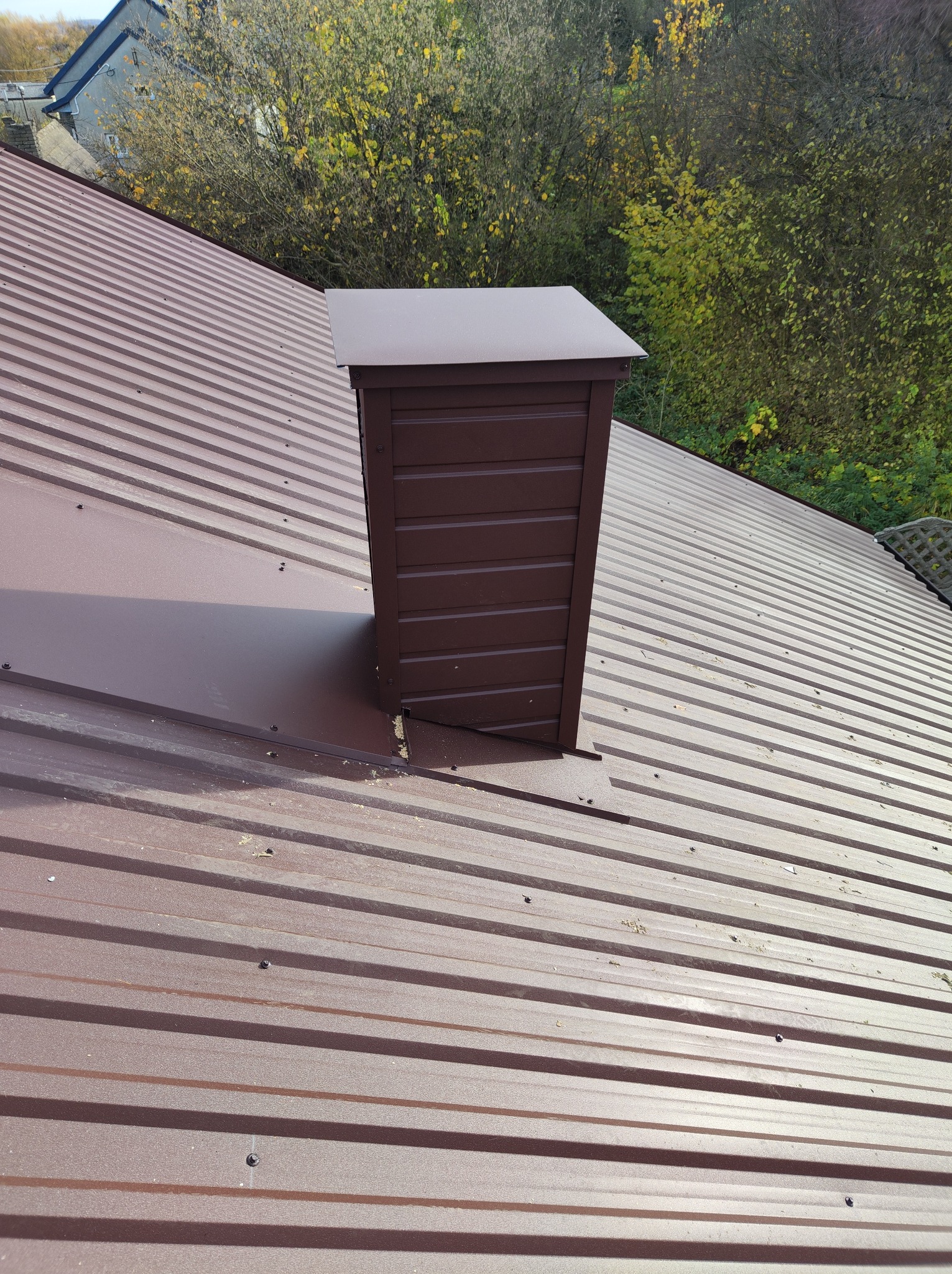 Ciemnobrązowy dach z blachy trapezowej z kominem wentylacyjnym, widok z góry, jesienne drzewa w tle.