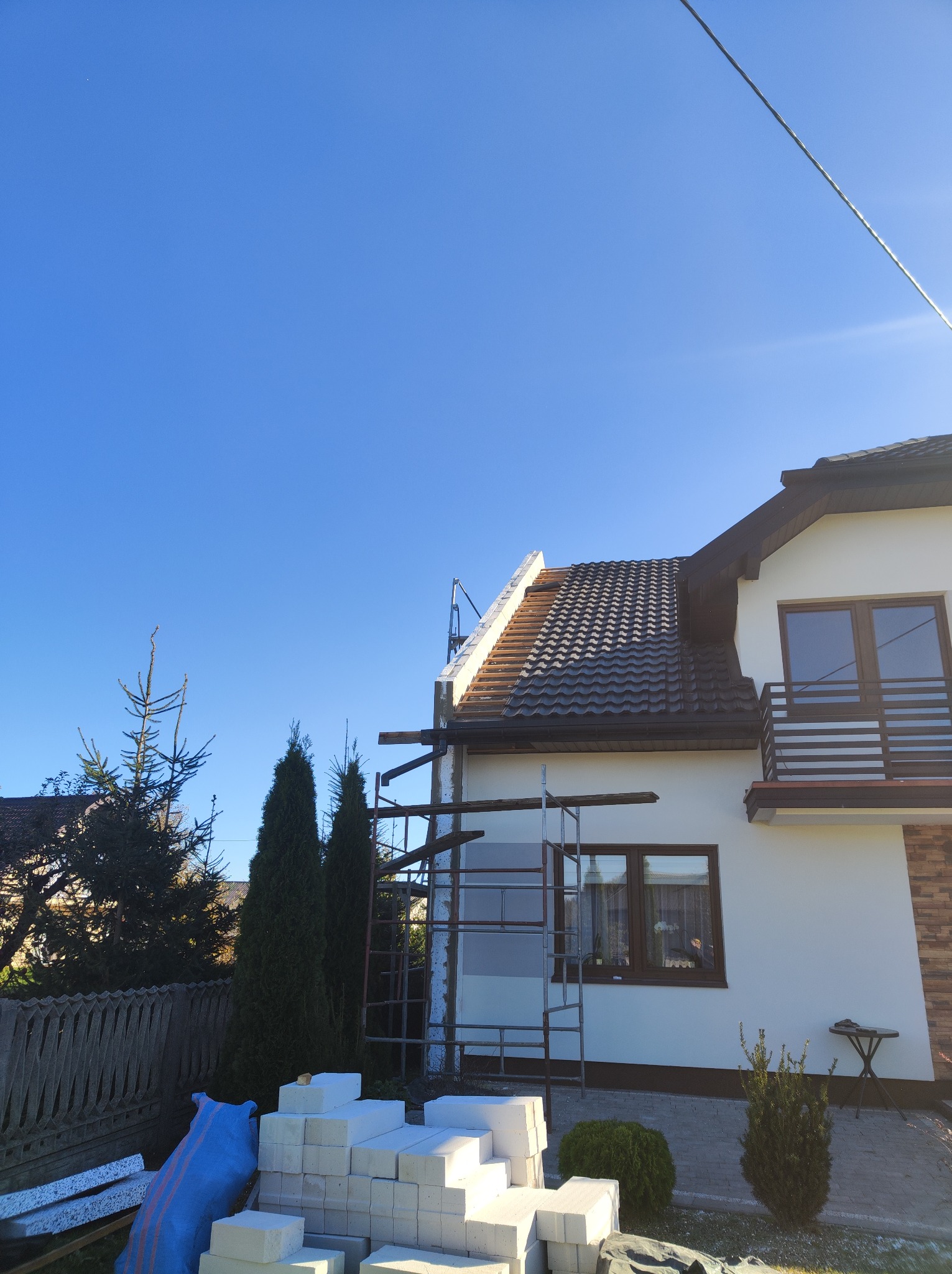 Dom jednorodzinny w trakcie prac dekarskich, widoczne rusztowania, częściowo zdjęte stare pokrycie dachu i ułożona nowa dachówka w kolorze grafitowym, na dole ułożone bloczki styropianowe.