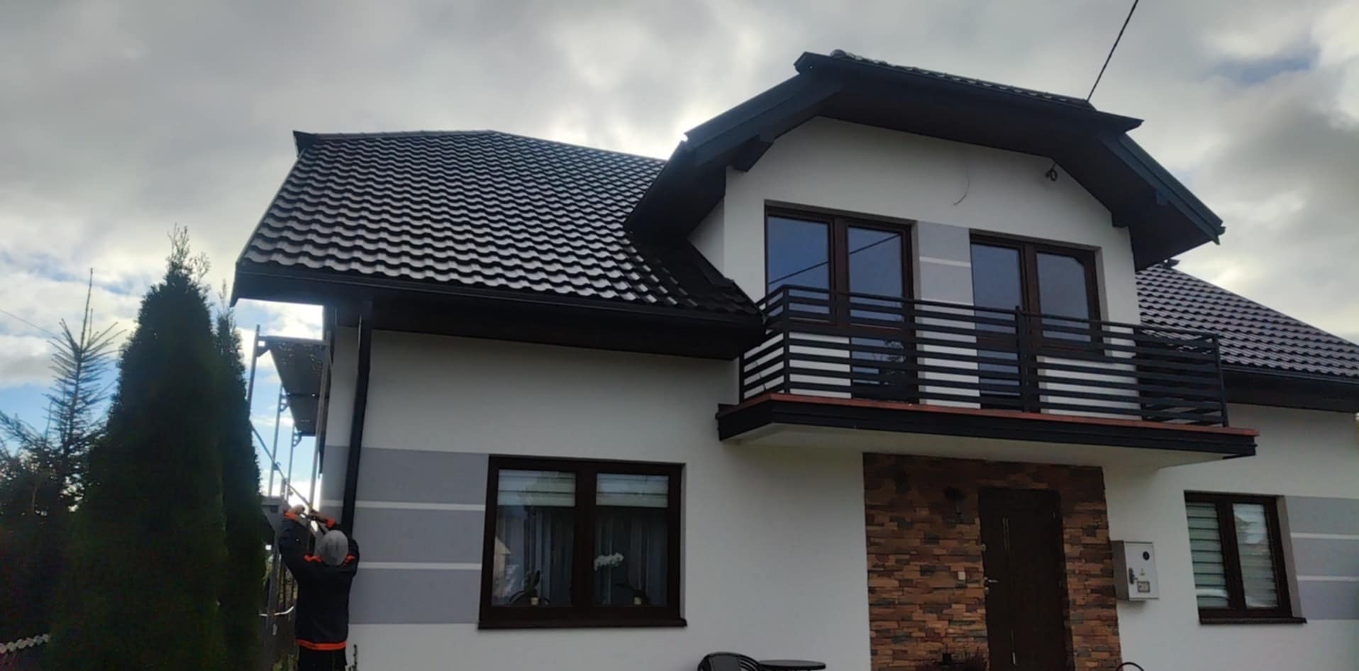 Pracownik na rusztowaniu montuje rynnę na tle domu z ciemną dachówką i balkonem.